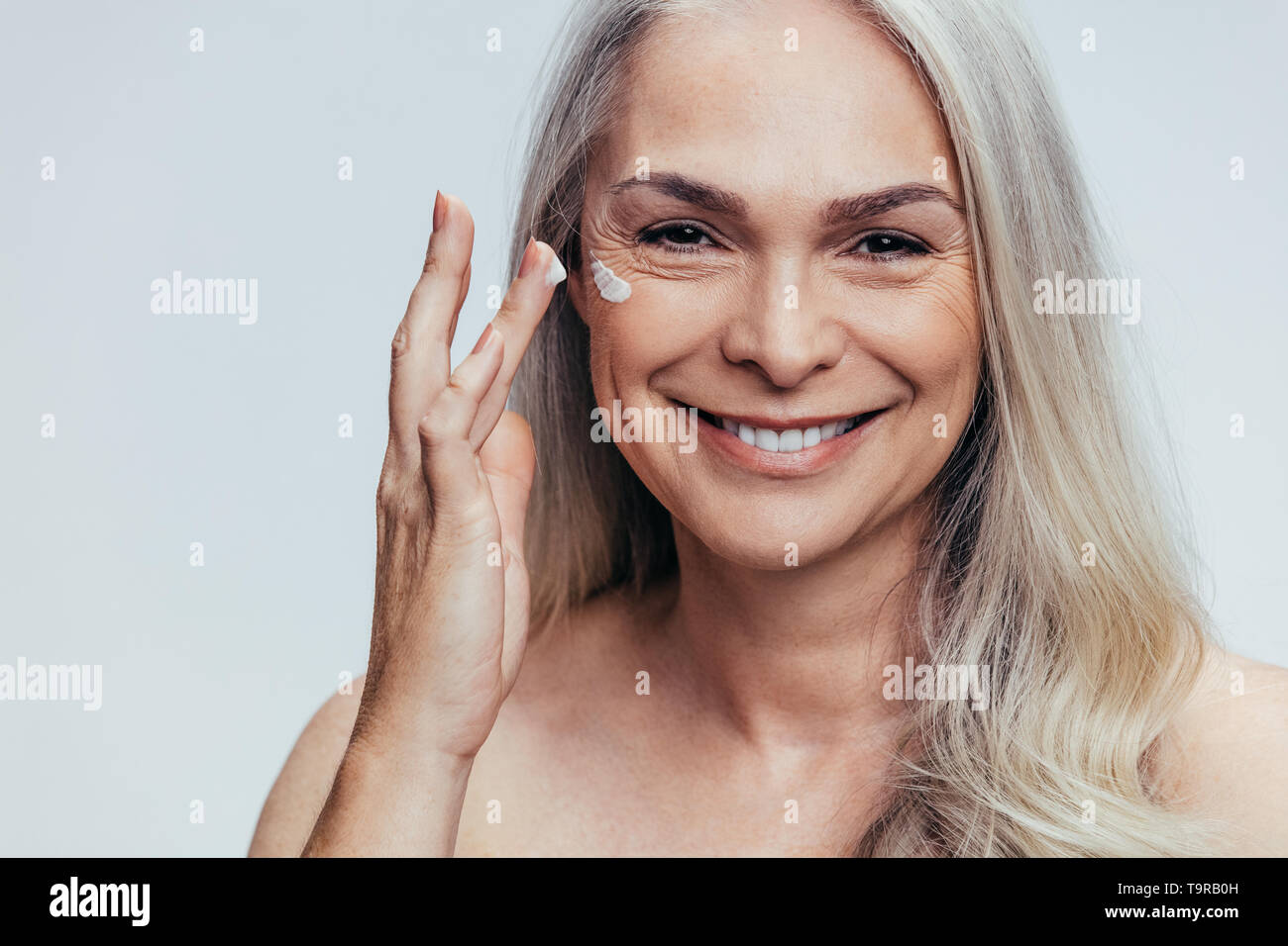 Smiling mid adult caucasian woman applying anti aging cream on her face. Senior female woman applying moisturizer on her face against grey background. Stock Photo