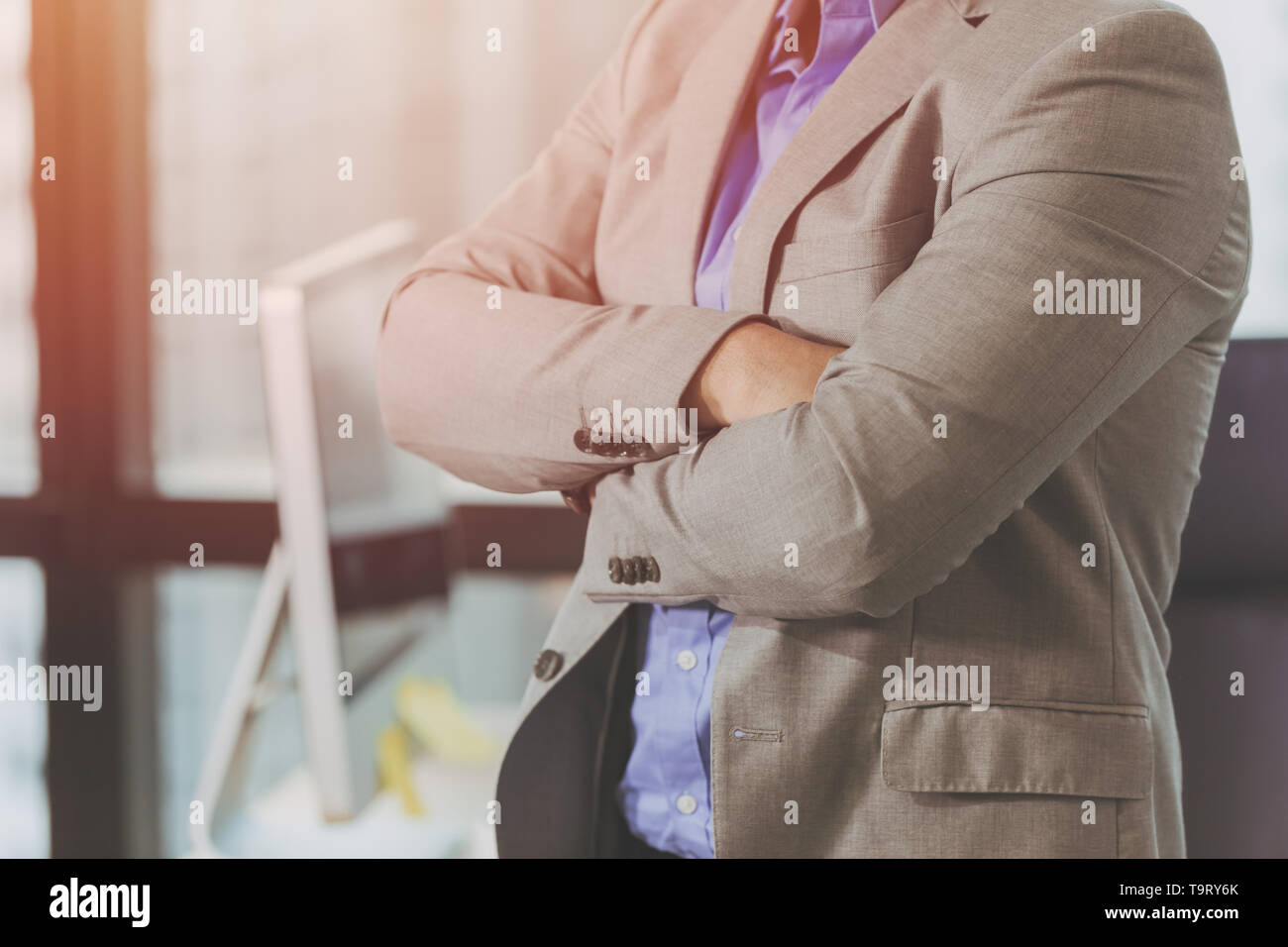 Confident businessman in suit standing with arms folded in business office background Stock Photo