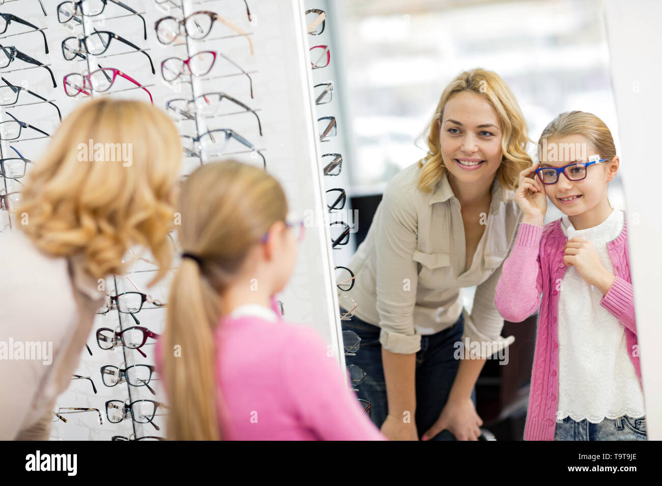 Female child look herself in mirror with blue frame for eyeglasses in optician shop Stock Photo