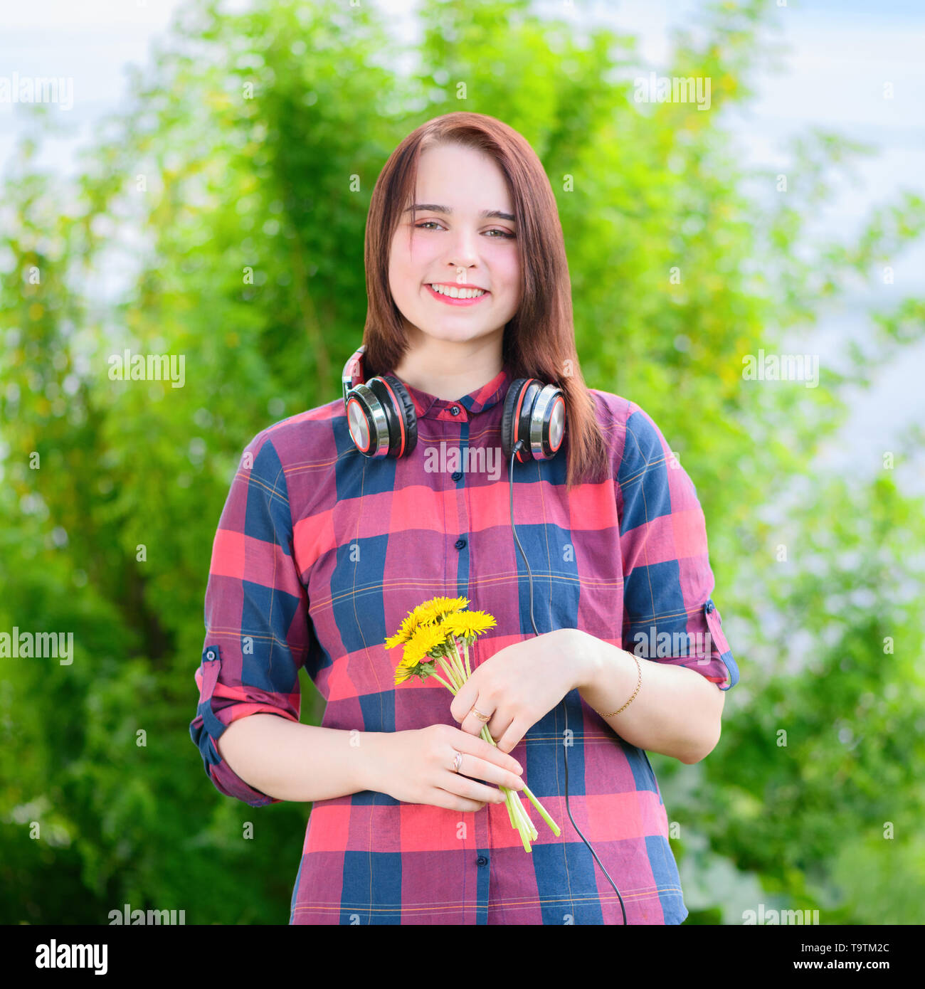 The smiling brunette woman is holding the bouquet of yellow flowers in her hand in a summer park. Stock Photo