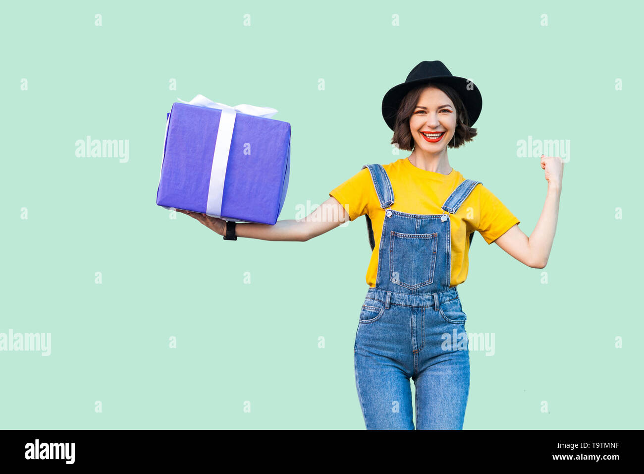 Happy attractive young girl in hipster wear in denim overalls and black hat standing and holding big heavy gift box with toothy smile, show strong arm Stock Photo