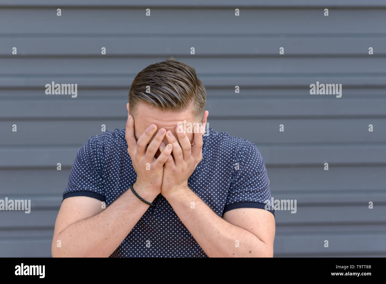 Depressed or despondent young man overcome by hopelessness standing with his head in his hands in front of a grey wall with copy space Stock Photo