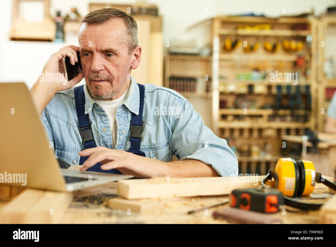 Portrait of modern senior carpenter speaking by phone and using laptop while working in joinery, copy space Stock Photo