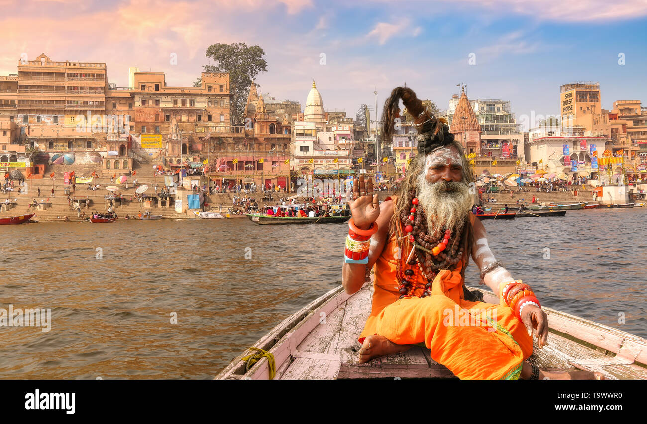 Sadhu baba sitting on a wooden boat overlooking historic Varanasi city architecture with Ganges river ghat at sunset. Stock Photo