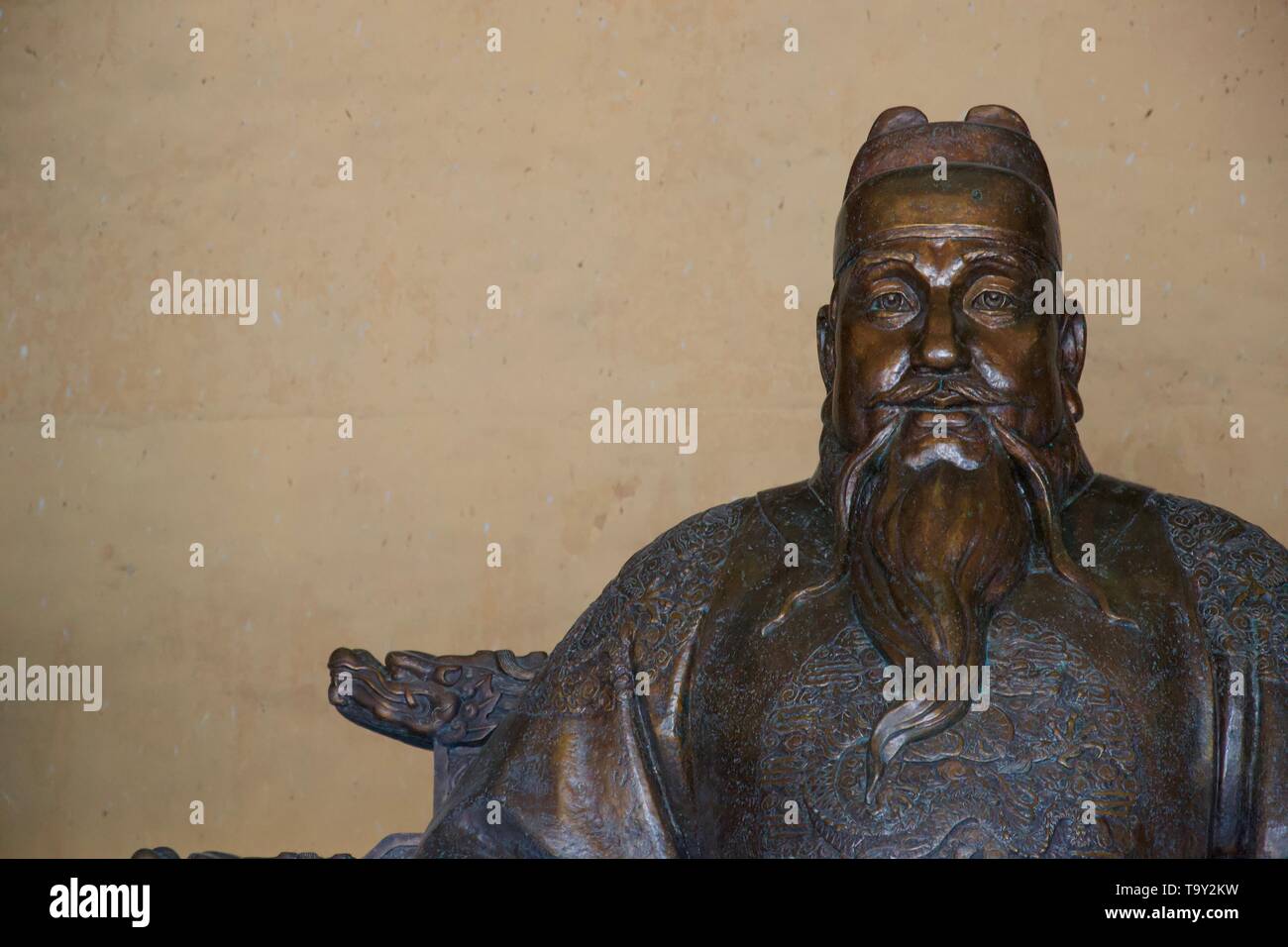 Statue of sad tired old Chinese man with big eyes, a whispy beard, ornately embroidered shirt and a hat that looks like a Lego brick. Made from a gold Stock Photo