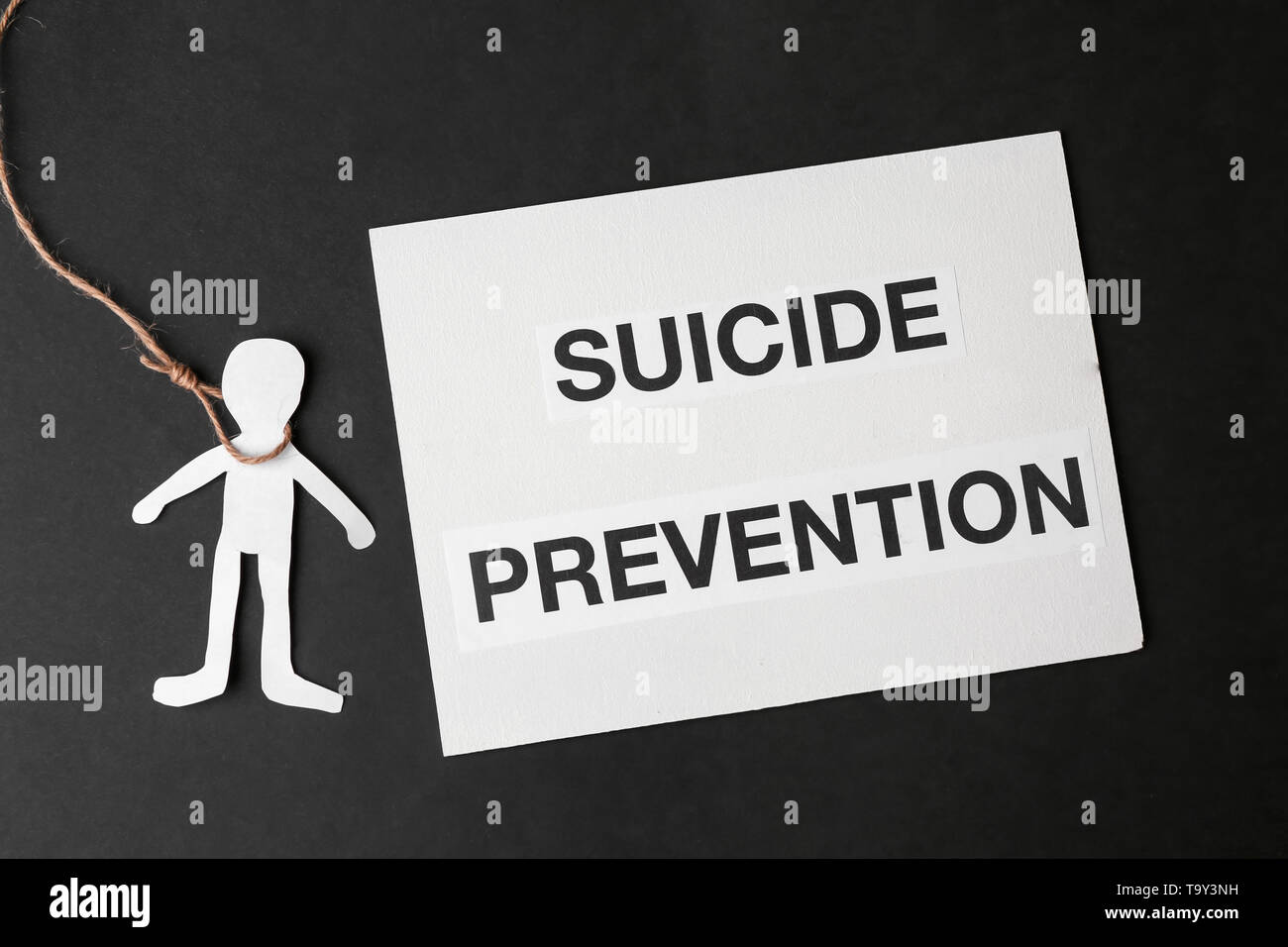 Paper human figure with noose around neck and sheet with text SUICIDE PREVENTION on dark background Stock Photo