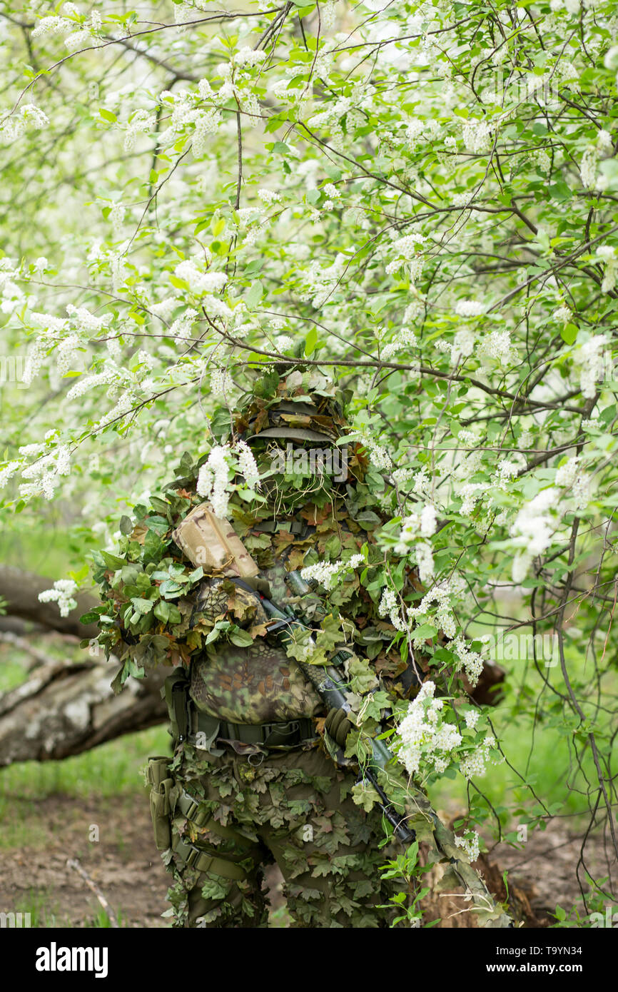 Cute portrait of a military male sniper in camouflage, mask, cap and rifle on his hands against the background of a flowering cherry in the summer out Stock Photo