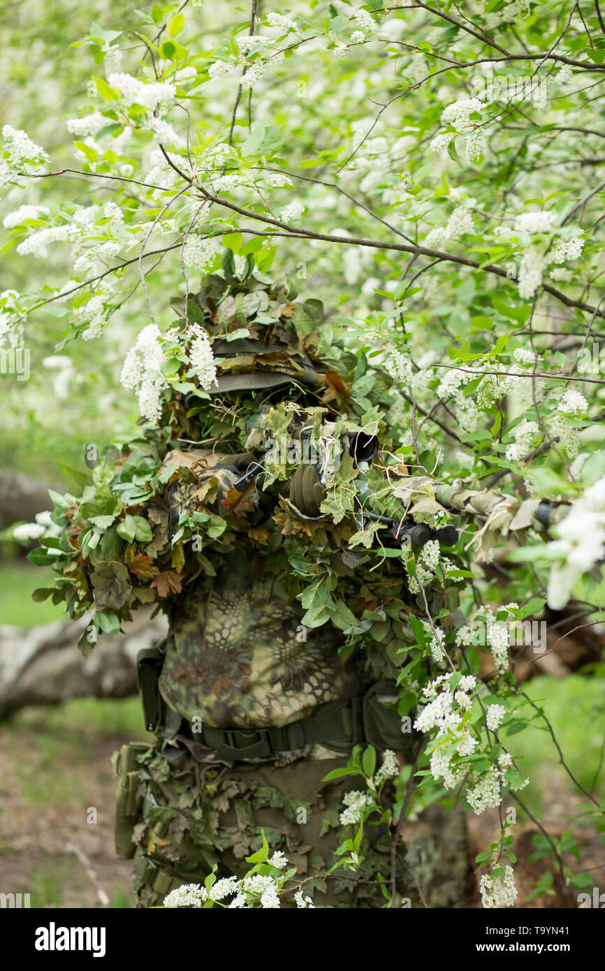 Spring portrait of a military sniper man in camouflage, mask, cap shooting at the enemy in the summer in nature near the white flowering trees. Army,  Stock Photo