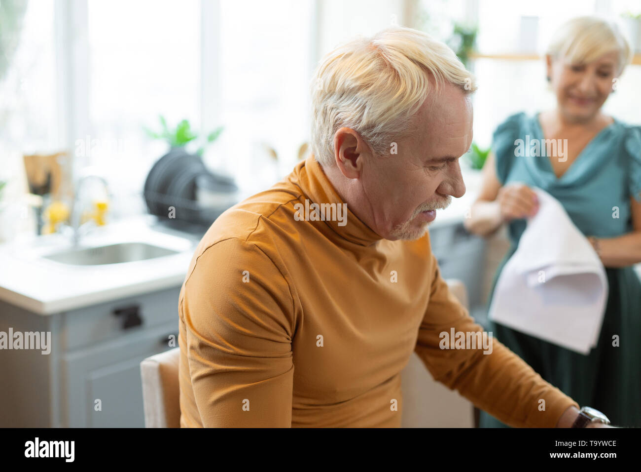 Stylish handsome attractive grey-haired bearded man sitting at the kitchen. Stock Photo