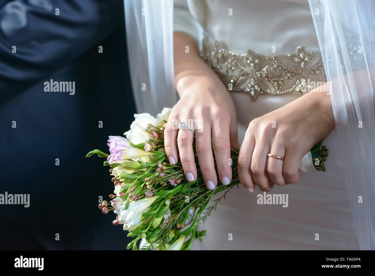 Bride and groom at the wedding with focus on bride's manicured hands with engagement ring and gold wedding band, holding a bohemian rustic bouquet Stock Photo