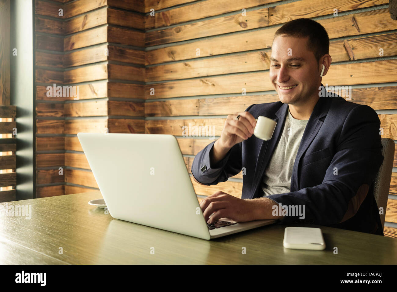 Smiling successful businessman drinking coffee in the office as he browses on an open laptop while wearing an audio earbud Stock Photo