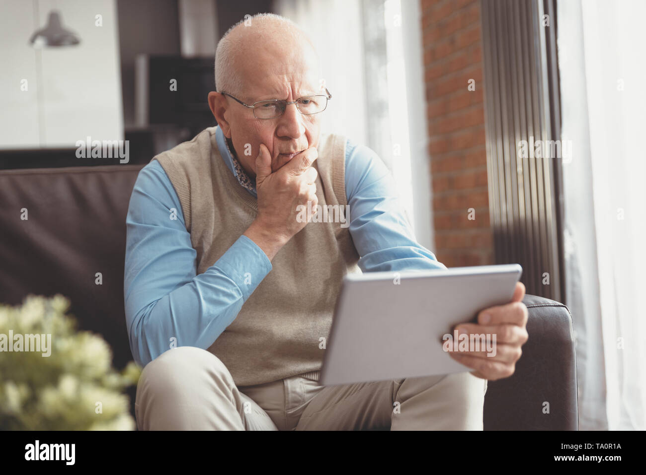 Senior man surprised using a digital tablet while sitting on couch at home Stock Photo