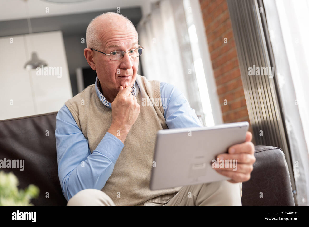 Senior man surprised using a digital tablet while sitting on couch at home Stock Photo