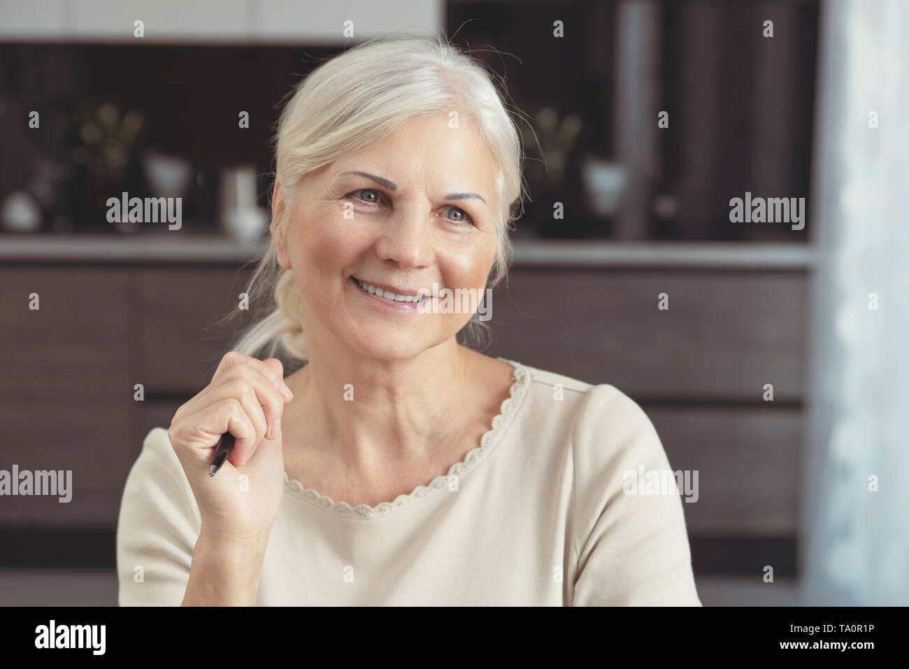 Cheerful senior woman portrait. Pensioner woman holding a pen, sits at home and looks at the window. Concept of a happy retirement Stock Photo