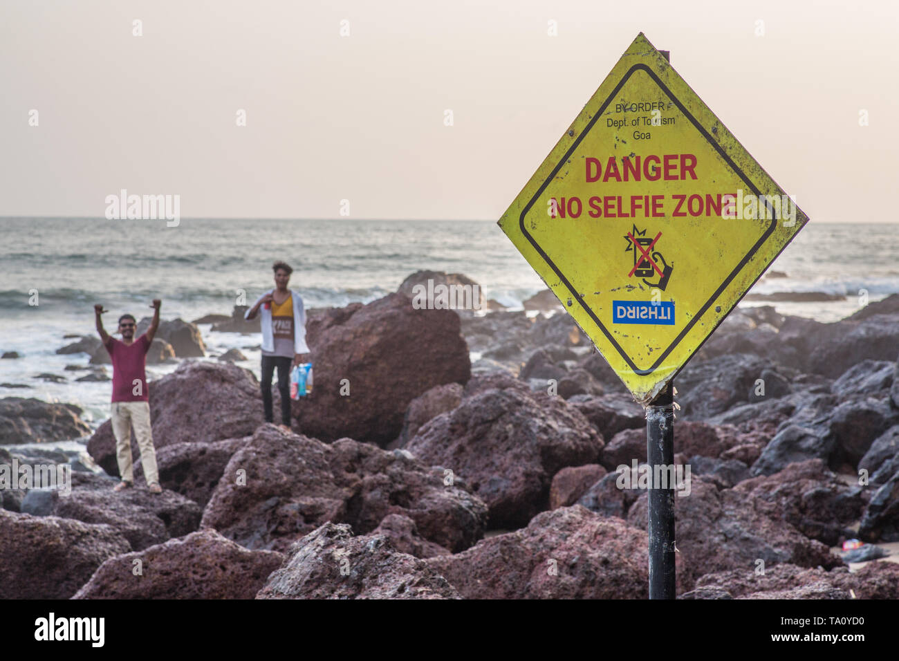 A sign warning visitors from shooting selfies on the rocks in this tropical location while people in the background are ignoring the same. Stock Photo