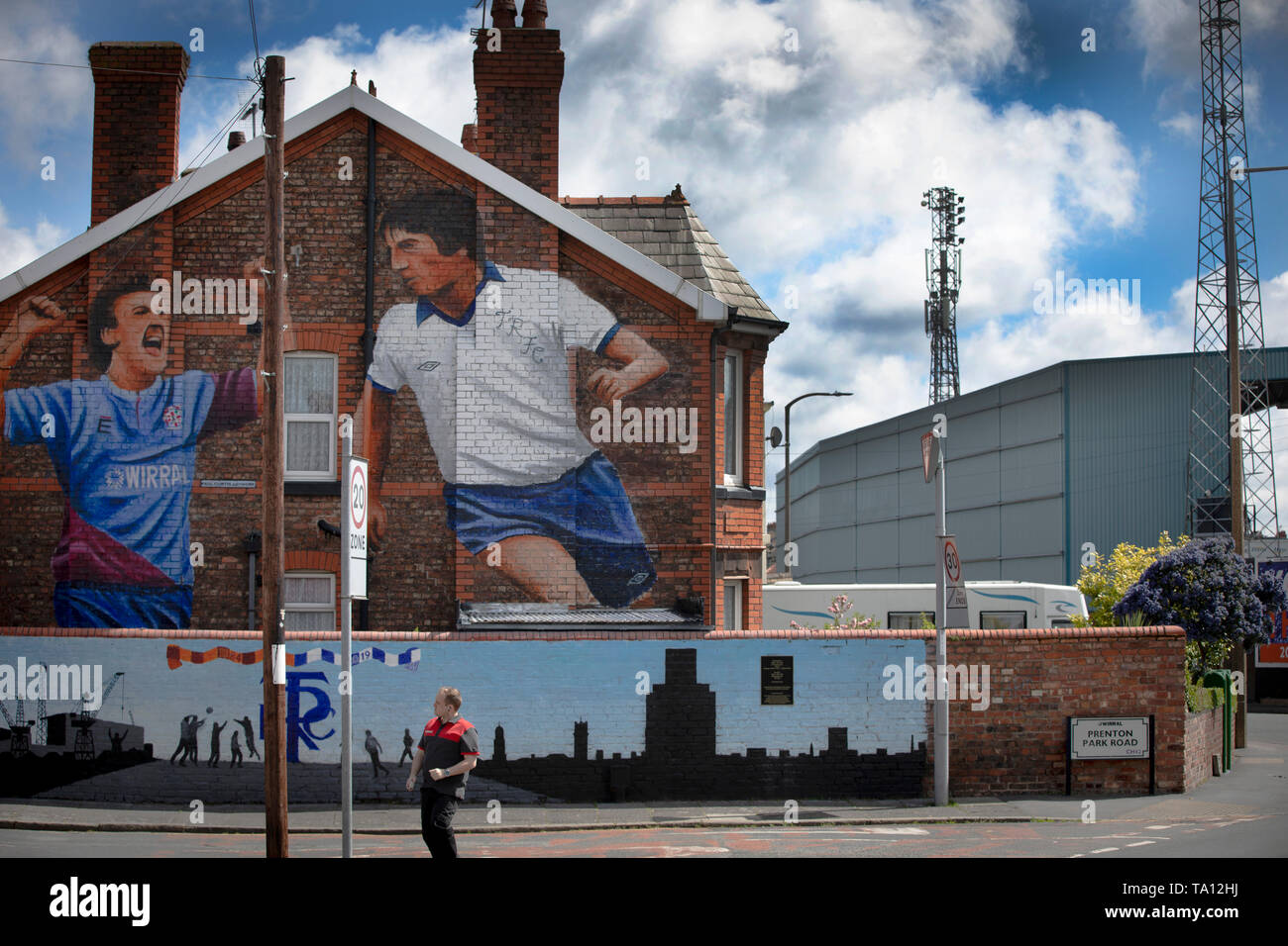 A man walking past a mural by artist Paul Curtis on a the gable end of a house in Prenton, Wirral depicting two Tranmere Rovers players. The mural is located just outside the club's stadium, Prenton Park. Tranmere Rovers gained promotion to EFL League 2 from the National League via a play-off in the 2017-8 season and returned to Wembley 12 months later on 25th May to face Newport County for a place in EFL League One. Stock Photo