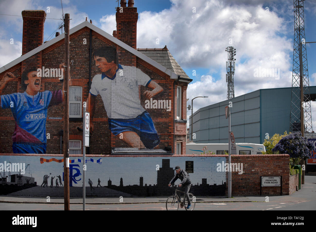 A man cycling past a mural by artist Paul Curtis on a the gable end of a house in Prenton, Wirral depicting two Tranmere Rovers players. The mural is located just outside the club's stadium, Prenton Park. Tranmere Rovers gained promotion to EFL League 2 from the National League via a play-off in the 2017-8 season and returned to Wembley 12 months later on 25th May to face Newport County for a place in EFL League One. Stock Photo
