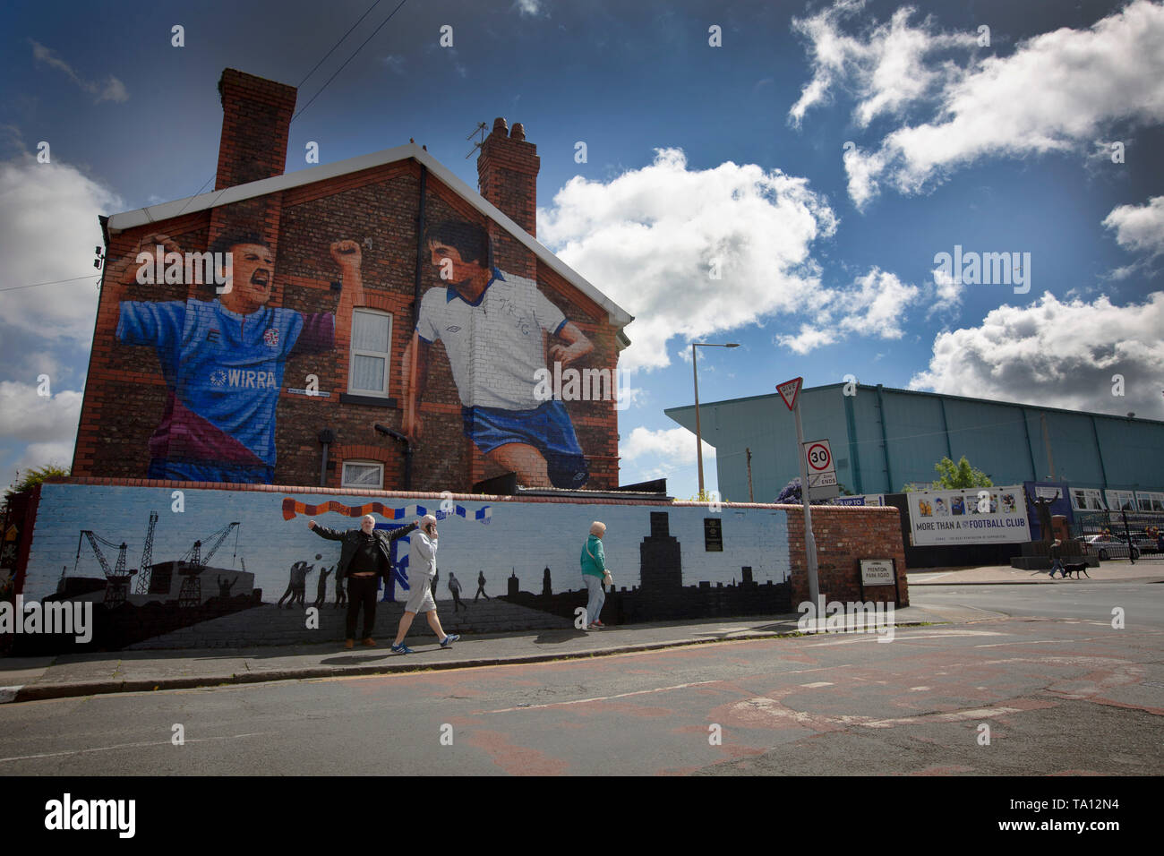 People walking past a mural by artist Paul Curtis on a the gable end of a house in Prenton, Wirral depicting two Tranmere Rovers players. The mural is located just outside the club's stadium, Prenton Park. Tranmere Rovers gained promotion to EFL League 2 from the National League via a play-off in the 2017-8 season and returned to Wembley 12 months later on 25th May to face Newport County for a place in EFL League One. Stock Photo