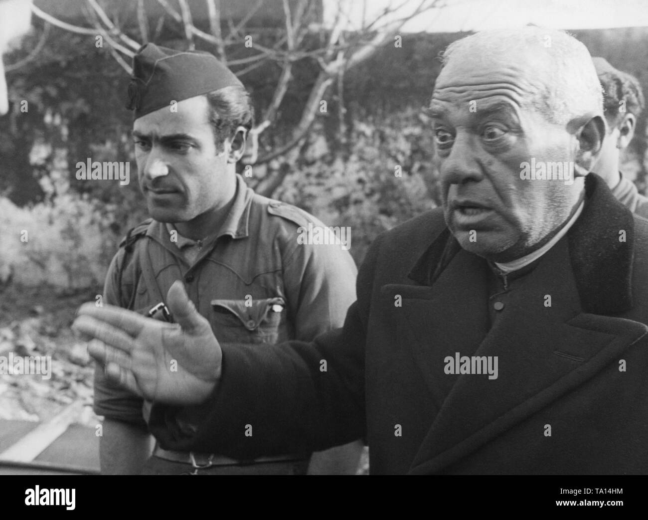 A Catholic Spanish priest in a black soutane and coat (right) tells Spanish soldiers something with eyes wide open. On the left, a French soldier of Franco's troops (with a Gorillo cap). Stock Photo