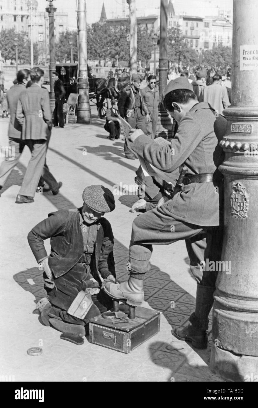 A Spanish national soldier lets a man clean his shoes, while he is reading a newspaper, in front of a lantern on the Rambla (promenade in the center) in Barcelona, ??Catalonia, Spain in March 1939, after the conquest of Barcelona by General Francisco Franco in January, 1939. In the background, flaneurs and soldiers. On the lantern, a call of Franco to the inhabitants of Barcelona. Down below on the cast-iron lantern,  the coat of arms of Barcelona. Stock Photo