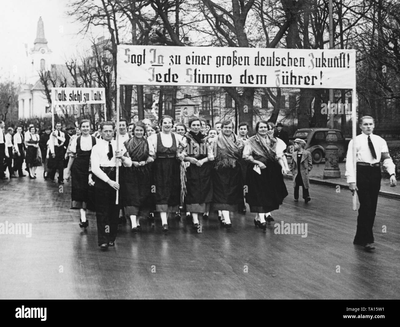 A group of Sudeten German women in traditional costumes on their way to the polling location. In the Sudeten German by-elections, votes are cast concerning the annexation of the Sudetenland to the German Reich. On the posters: 'Say Yes to a great German future! Everybody vote for the Fuehrer! ' and 'People stand by people!'. Stock Photo