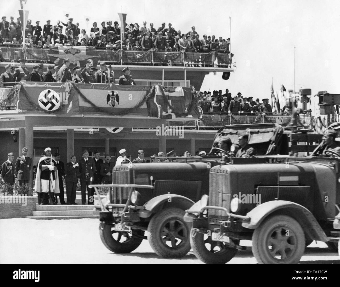 Photo of German military vehicles of the Condor Legion during a victory parade for General Francisco Franco at Madrid Barajas airport, after the invasion of the city on March 28, 1939. In the background, the terminal of the airport, which was turned into a tribune, with the VIP stand for Franco (salutes with raised arm). Beneath it, a swastika flag and the tricolor (flag of the Kingdom of Italy) are hanging next to the Spanish Bandera. Stock Photo