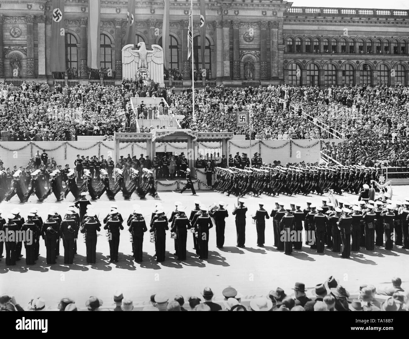 Photo of the VIP stand during the parade for the Condor Legion on the East-West Axis (former Chalottenburger Chaussee, today Strasse des 17. Juni) in front of the main facade of the Technische Universitaet in Berlin on June 6th, 1939. Adolf Hitler (under a canopy) is giving the marching soldiers the Nazi salute. On the left under the Reichsadler (imperial eagle) with a swastika, a radio commentator. The music band of the Kriegsmarine (war navy) is playing. Stock Photo