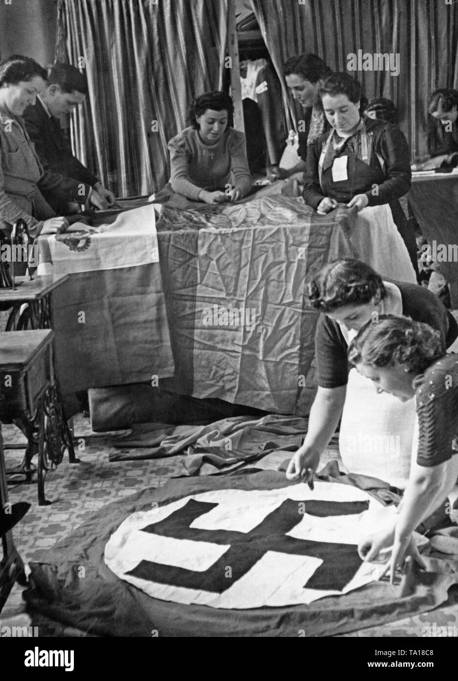 Undated photo of a group of Spanish women sewing Spanish, Italian and German flags for the Condor Legion in a workshop. In the foreground two women are sewing a swastika onto a flag. On the table, two banderas are being sewn. Stock Photo