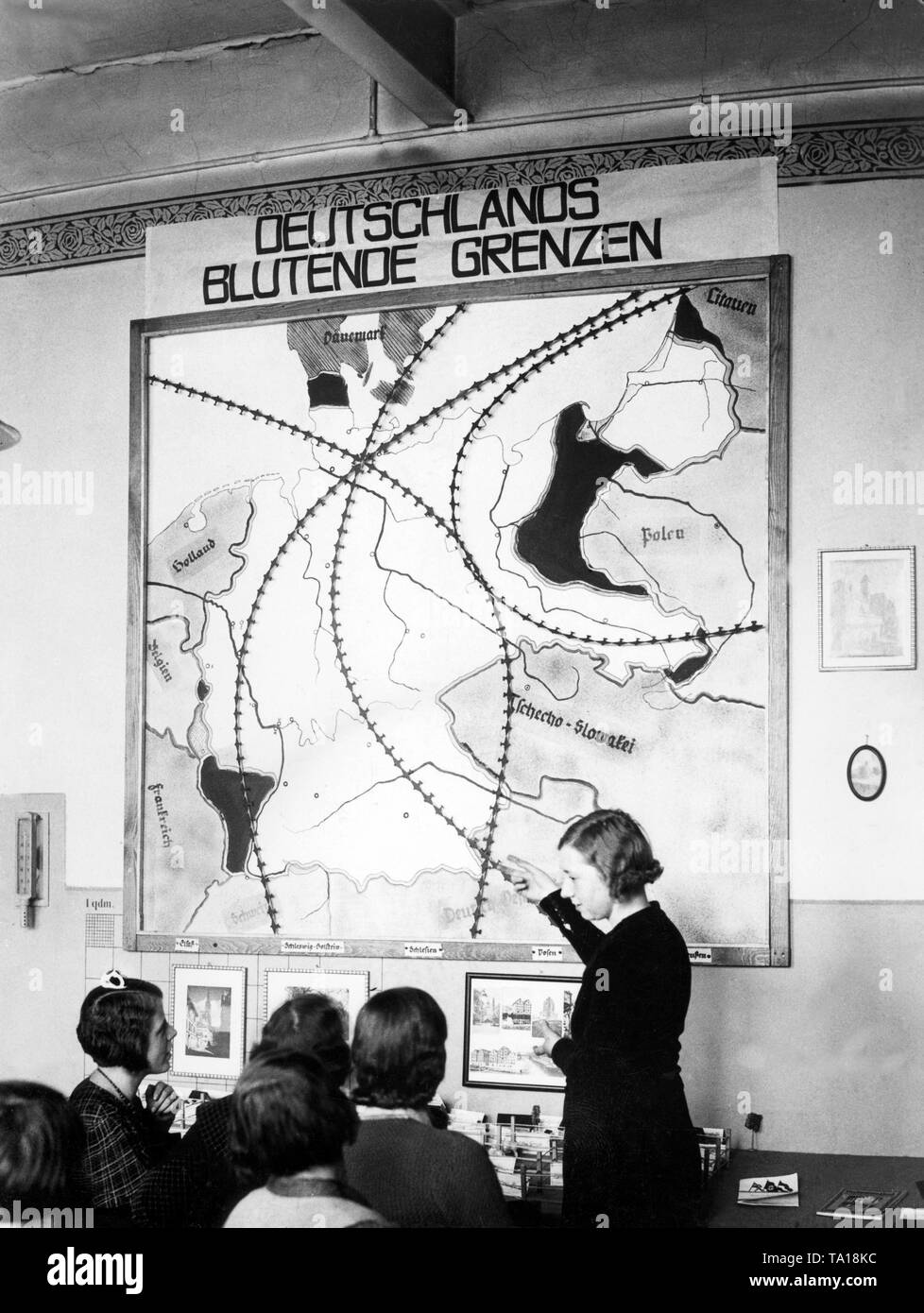 On the map entitled "Germany's Bleeding Boundaries" not only the lost  territories of Germany are depicted, but also how deeply hostile airplanes  can penetrate into the imperial territory. A teacher points to