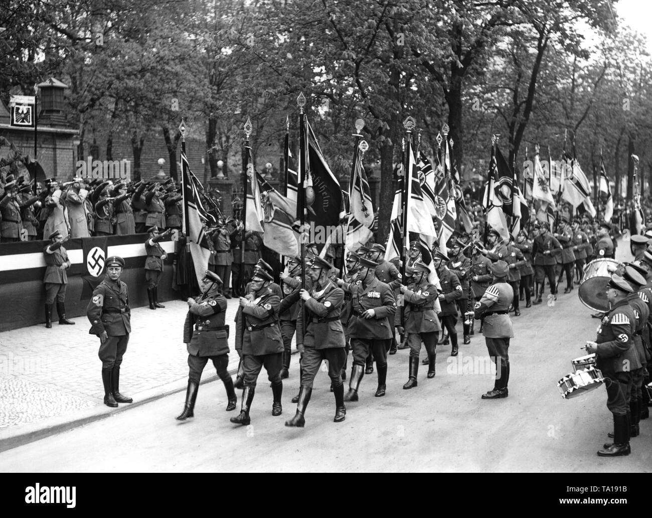 Photo of the flag parade of the National Socialist Alliance of Red Front-Fighters as it passes by General Ewald von Lochow (left, on the VIP stand with Pickelhaube) at the Jahnsportplatz in the Hasenheide, Berlin.The NS Alliance of Red Front-Fighters present their new flags, after the black ribbons have been abolished in memory of the Versailles Treaty due to the reintroduction of military service. To the right, a music band. Stock Photo