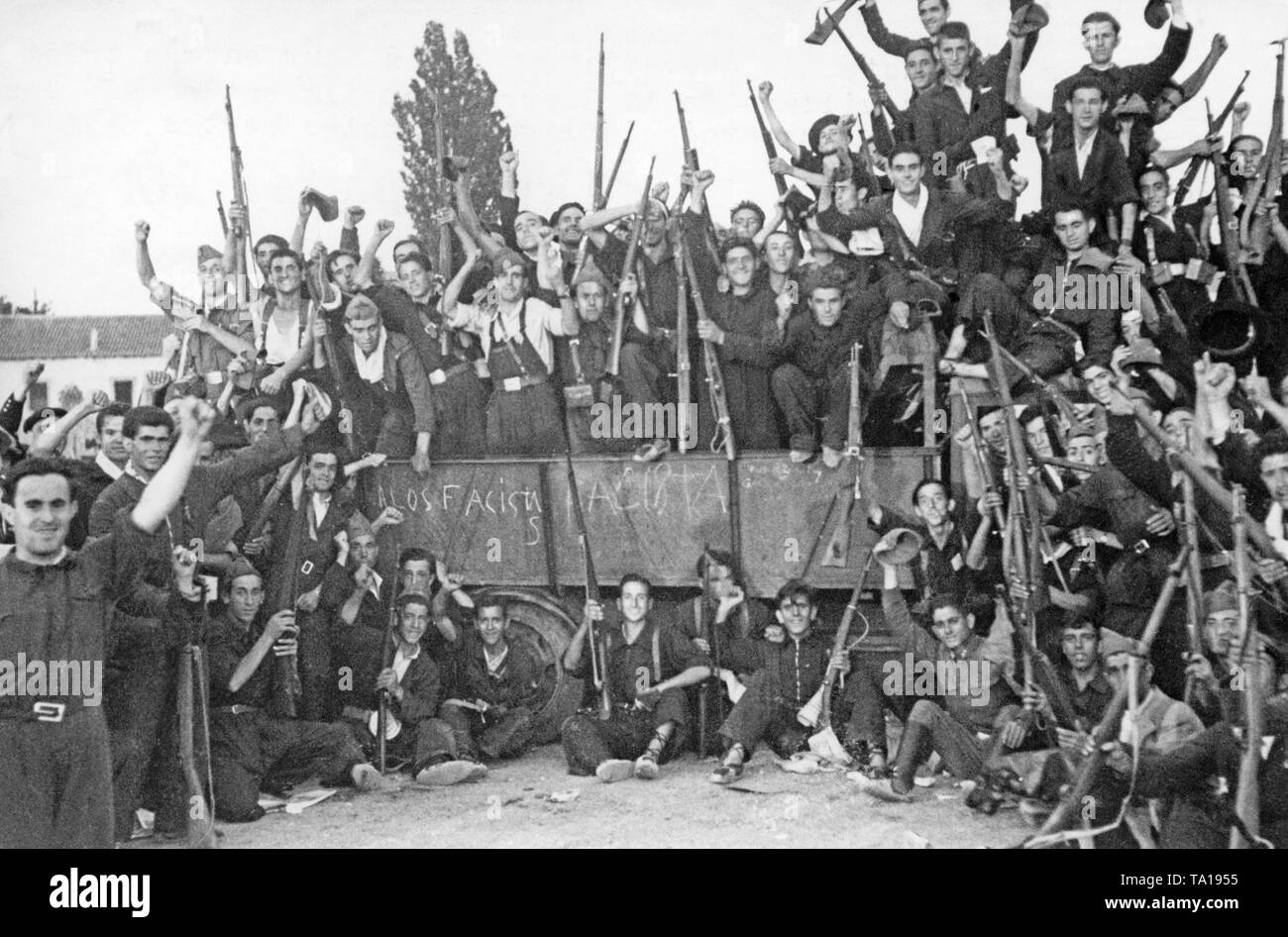 Photo of a cheering group of Republican fighters, presumably anarchist (FAI, CNT) volunteers in blue overalls in front of a truck. The fighters are armed with rifles and wear simple shoes, mostly alpargatas, simple cotton shoes with braided hemp soles. On the planks of the truck: 'A los Fascistas' (Against the fascists). Stock Photo