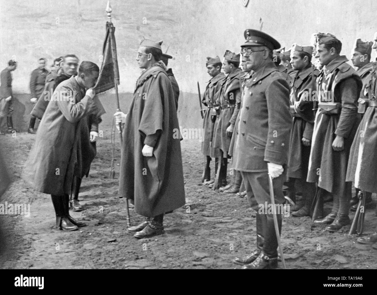 The photo shows an officer in uniform and loden cape as he kisses the Bandera (the red-yellow-red flag of Spain, held by a major) during his swearing-in. Previously, there was a worship service in the Burgos Officer Candidate School in Castile and Leon, Spain. In the front, an instructor and a lieutenant with a saber and an umbrella. Stock Photo
