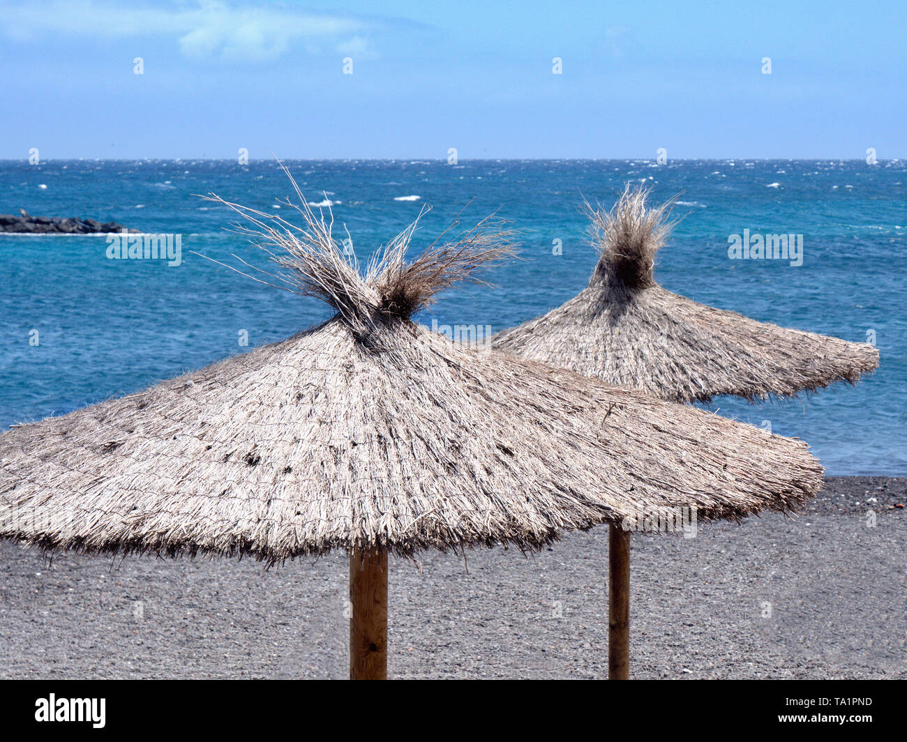 in front of the dark blue atlantic ocean, two reet grass parasols stand by the water on a dark volcanic beach in close-up.over it dark blue sky, pictu Stock Photo