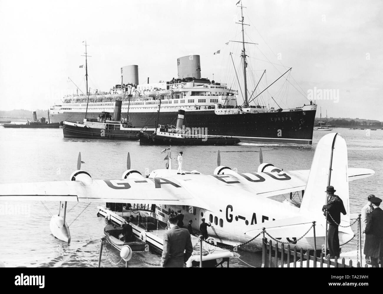 The fast steamer 'Europa' of Norddeutscher Lloyd is in the port of Southampton. In the foreground, the flying boat Short Empire G-AEUG Coogee of the Imperial Airways. On the left side of the flying boat is a raft, over which passengers can board the plane. Stock Photo