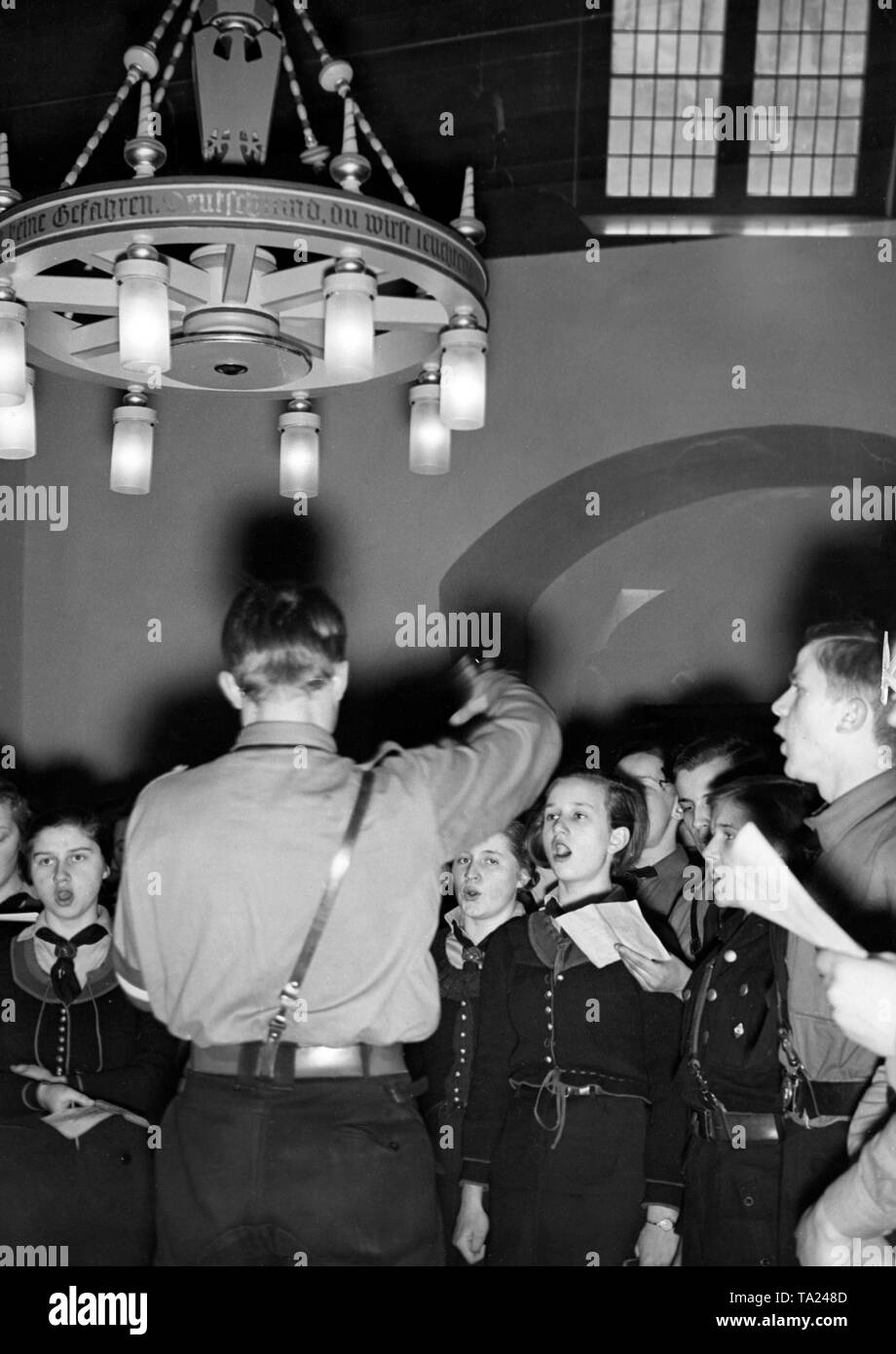 Members of the Hitlerjugend (Deutsches Jungvolk) as well as of the BDM are singing youth songs in the knights's hall of Storkow Castle. On the chandelier it says '... Youth is above the fear of danger. Germany you will shine ... 'from the Hitler Youth song' Our flag is fluttering us onward.' Stock Photo