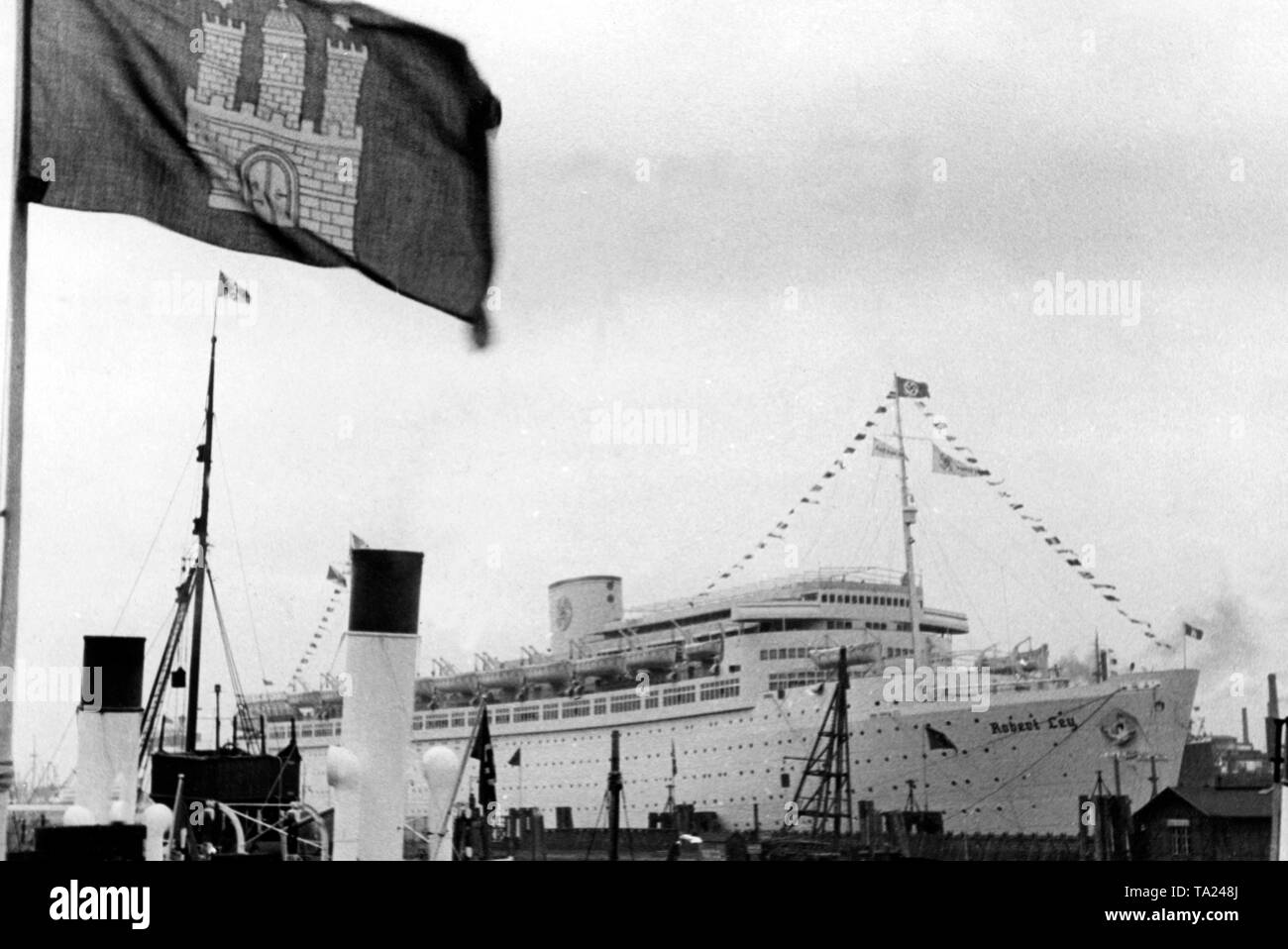 View of the cruise ship 'Robert Ley' of the Nazi organization 'Kraft durch Freude' ('Strength through Joy') in the port of Hamburg. In the foreground a flag with the coat of arms of Hamburg. Stock Photo