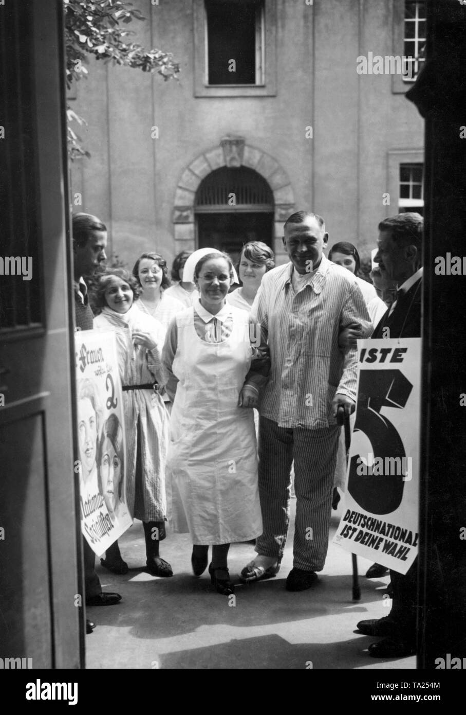 Patient is led to vote by a nurse at the Rudolf-Virchow hospital. On the left there is a supporter of the National Socialists with an election poster. On the opposite side stands a supporter of the German National People's Party with an election poster on which says 'German National is your choice' On the left, a poster of the NSDAP. Stock Photo