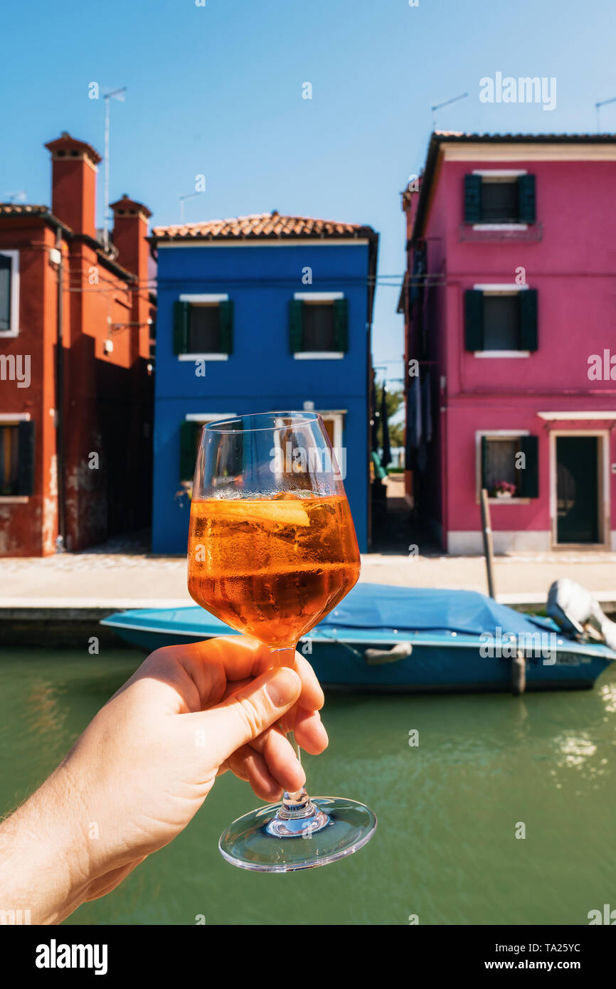 Hand of male holds glass with Spritz aperol cocktail overlooking colorful houses in Burano, Veneto, Italy Stock Photo