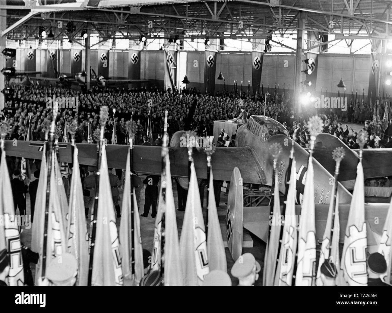 Rudolf Hess holds a speech at a conference of the Reich Chamber of Labor in the factory of Messerschmitt AG Haunstetten / Augsburg, and names the companies honored by Hitler, to which they awarded the title of 'National Socialist Model of Industry'. In the foreground, flags of the German Labor Front (DAF). Behind the lectern, a Messerschmitt Bf 110. Stock Photo