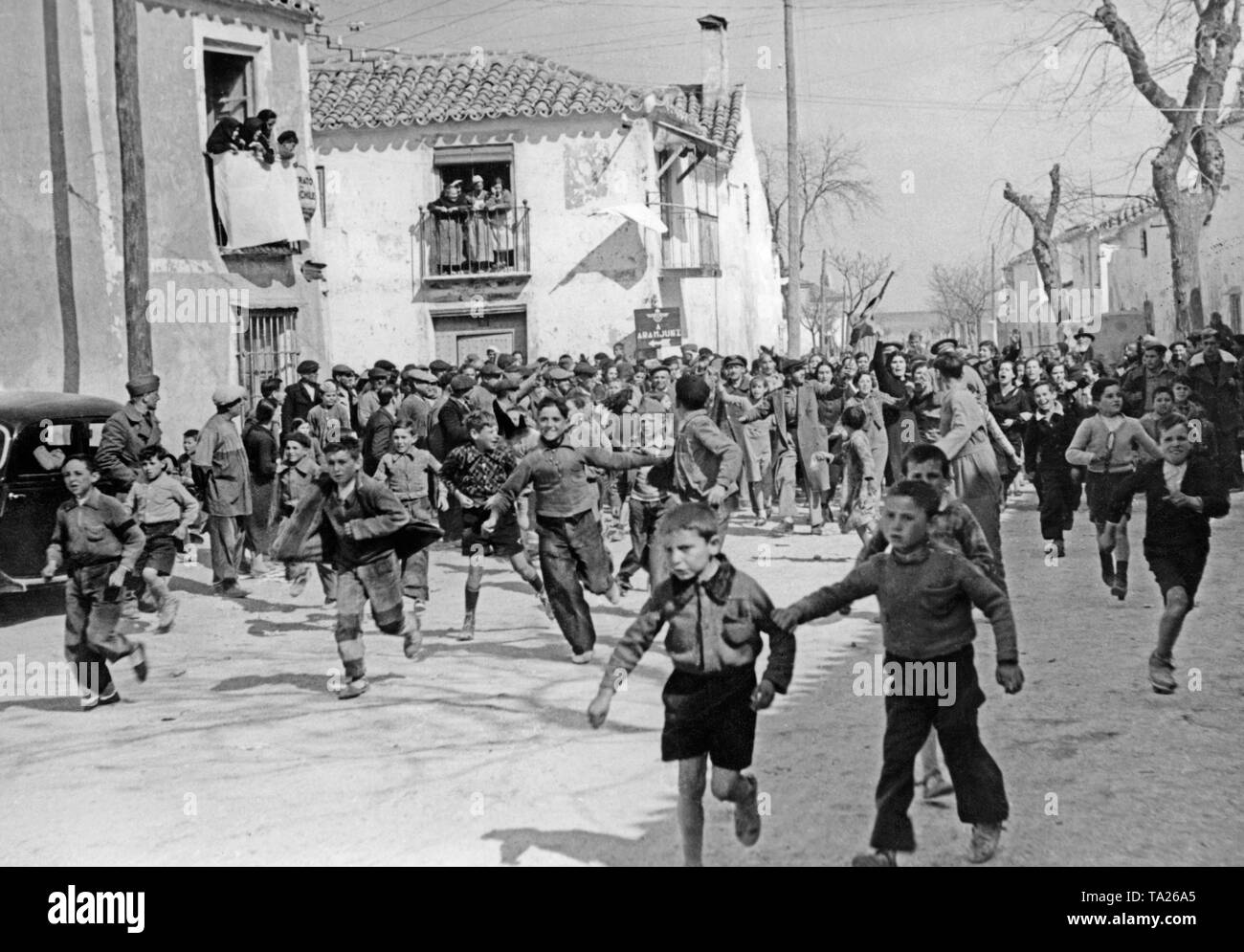 Photo of children and adults in the village of Templique near Madrid, who welcome the Spanish national troops under General Francisco Franco cheerfully. People swing the Bandera, the flag of the Nationalists. On the left on the houses there are white flags (sign of capitulation). Stock Photo