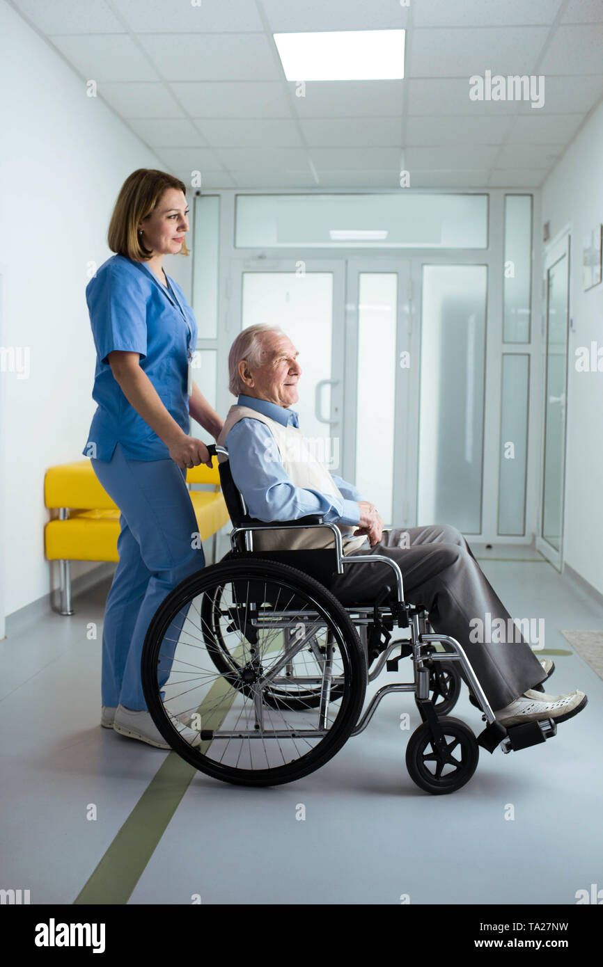 friendly nurse pushing senior patient in wheelchair at hospital corridor Stock Photo