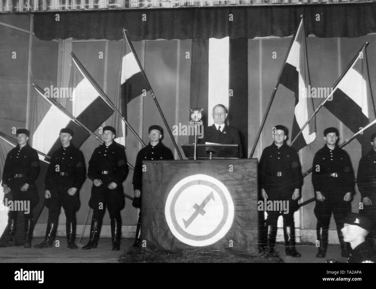 Dr. Ernst Neumann, chairman of the Memeldeutsche Kulturverband (symbol: Wolfsangel), gave a speech in the city of Memel (Klaipeda) to Memel German election workers on the state elections in the Memel area that will take place on 11 December 1938. Next to the lectern: Memeldeutscher Ordnungsdienst (Memel German Security Service) (SS). In the background, several Reich's flag. Stock Photo