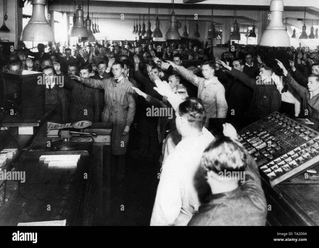 Employees sing the Horst Wssel Lied in the Scherl printing house at the end of the community reception. They perform the Nazi salute. On the right, a shadowbox, also called composing rack. A Volksempfaenger (radio) stands in front of the factory employees. Stock Photo