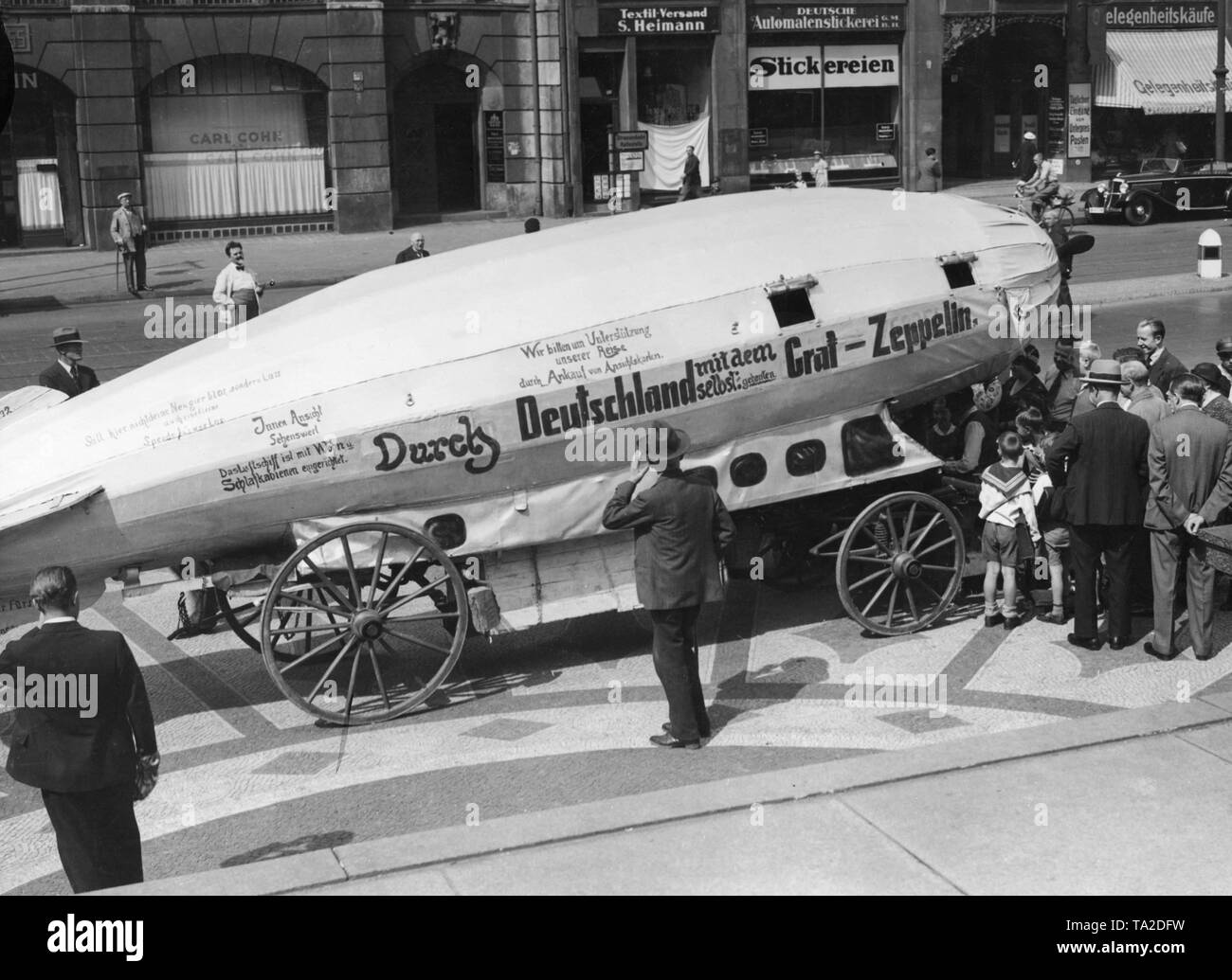 Unemployed people travel through Germany with a self-made zeppelin. The airship with a swastika pennant on the hull is viewed by passers at Hackescher Markt in Berlin-Mitte. In the background are the shops 'Carl Cohn A.G.', 'Textil-Versand S. Heimann', 'Deutsche Automatenstickerei' and 'Gelegenheitseinkaeufe'. Stock Photo