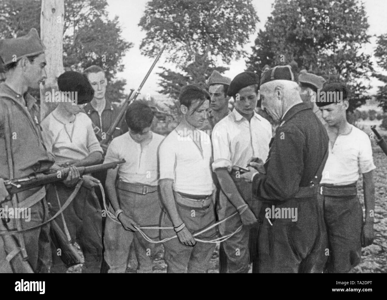 Exhausted young Republican prisoners are interrogated by Spanish soldiers (on the right, an armed falangist in a blue shirt, on the left, a soldier) tied with a rope. The soldiers (presumably Communists) wear bright shirts, breeches and berets. Stock Photo