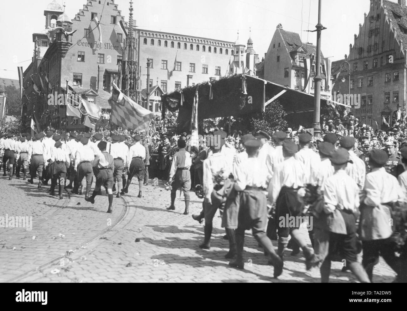 National youth organizations march in Nuremberg with Reichskriegsflagge (imperial war flag) and the Bavarian flag. Officers and generals are watching them in front of grandstands decorated with garlands. Stock Photo