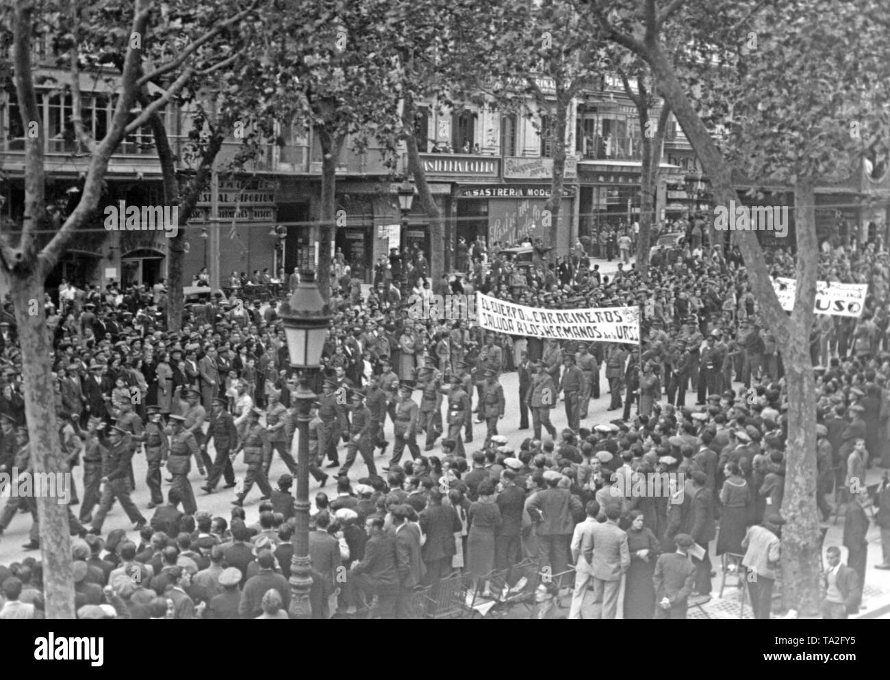 Photo of a Republican demonstration in remembrance of the 19. anniversary of the October Revolution (November 1917) on the Rambla (Promenade Las Ramblas) at the beginning of November, 1936. The marching military policemen carry a banner: 'The military police (Carabineros) greet their Soviet-Russian brothers.' The police officers in uniform raise their fists saluting. Passers-by watch the scene on the pavement. Stock Photo