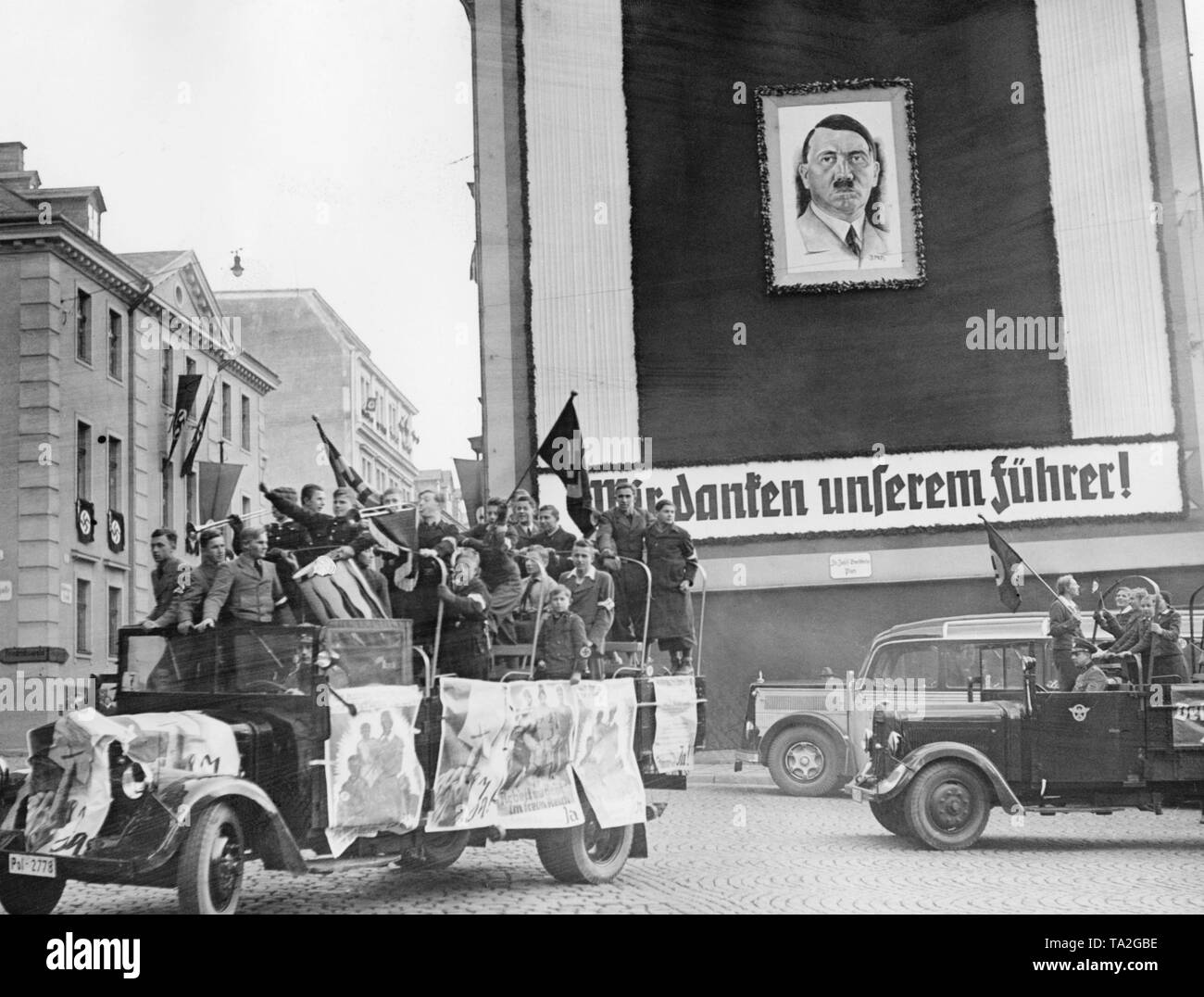 Trucks with members of the Deutsches Jungvolk (German Youth) drive through the street of a Sudeten German city. Election posters are attached to the truck. In the Sudeten German by-elections, votes are being cast concerning the annexation of the Sudetenland to the German Reich. In the background, a poster with a portrait of Adolf Hitler. Subtitle: 'We thank our Fuehrer!'. Stock Photo