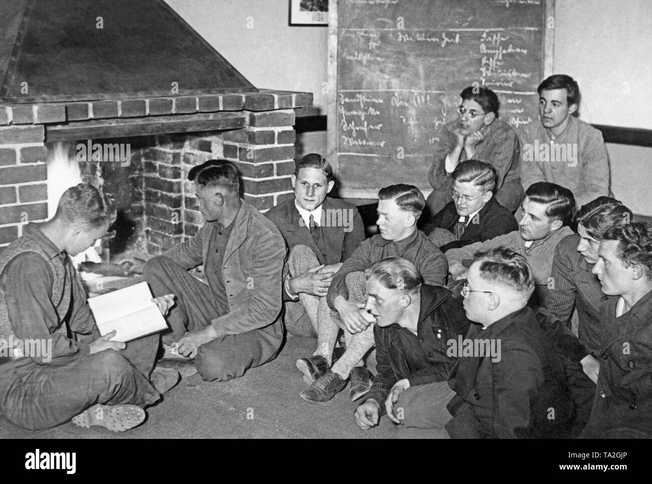 Members of the Deutsche Freischar sit together in front of a fireplace, one of the young men is reading from a book. Stock Photo