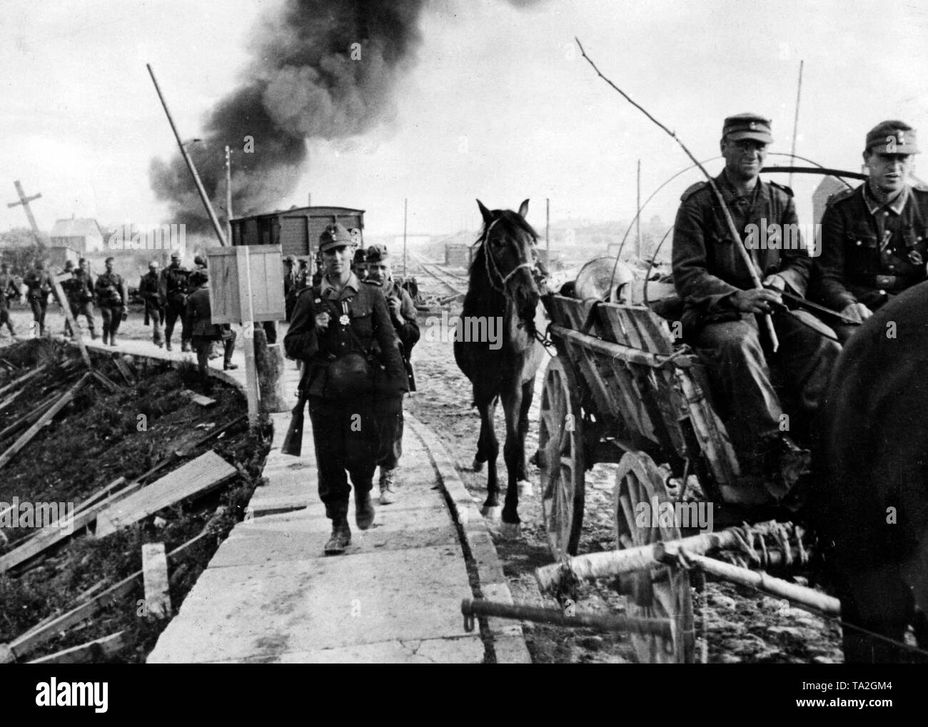 German soldiers on the return march in Latvia. In front, the group leader awarded with an Iron Cross 2nd class. In the background, a burning hut. Photo of the Propaganda Company (PK): war correspondent Schroeter. Stock Photo